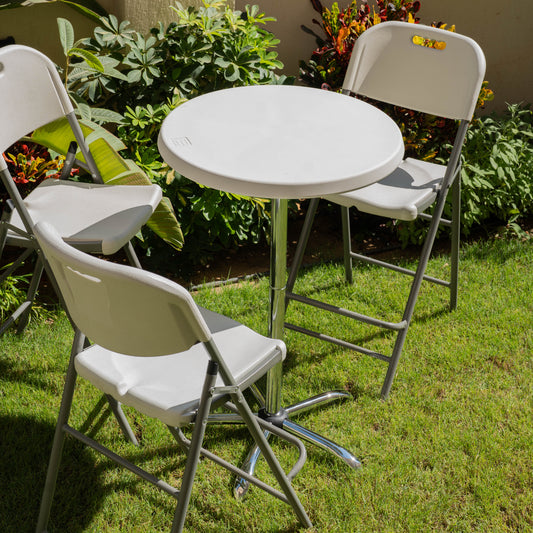 White folding table and chairs on grass with plants in the background