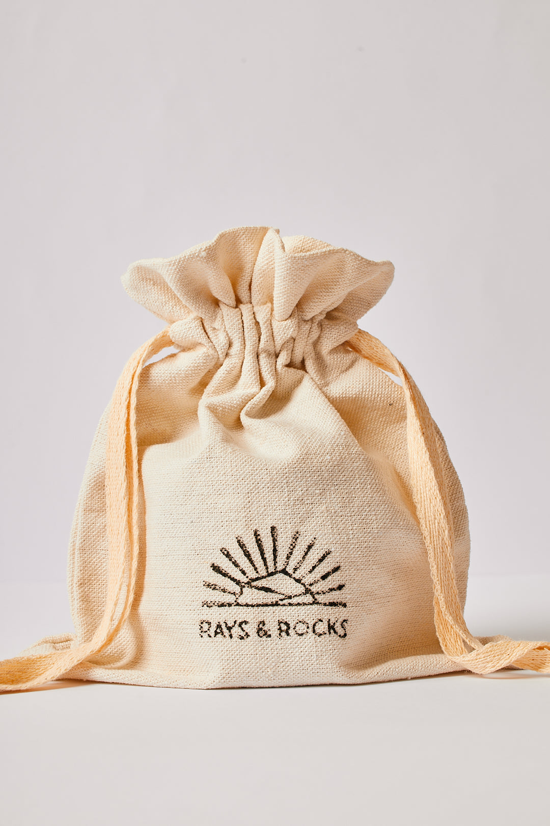 A beige handmade dust bag with a drawstring closure, featuring rays & rocks brand logo.